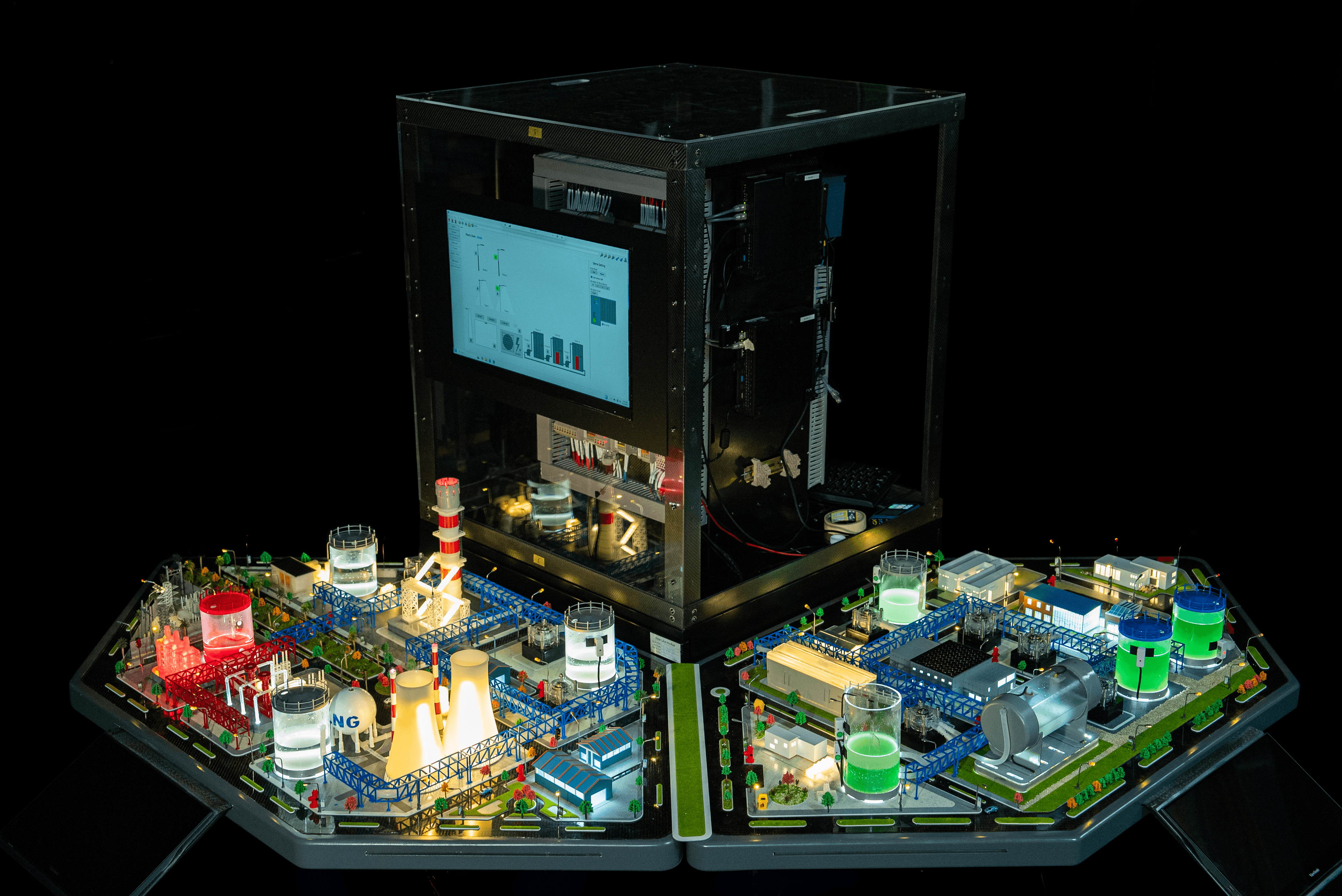 The control cabinet driving the range — an HMI screen and real PLCs racked behind glass, standing between two process panels with lit tanks, cooling towers and an LNG sphere